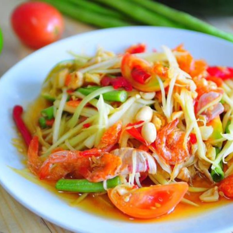Som Tum, a Thai papaya salad with shrimp, tomatoes, green beans, and peanuts on a white plate.