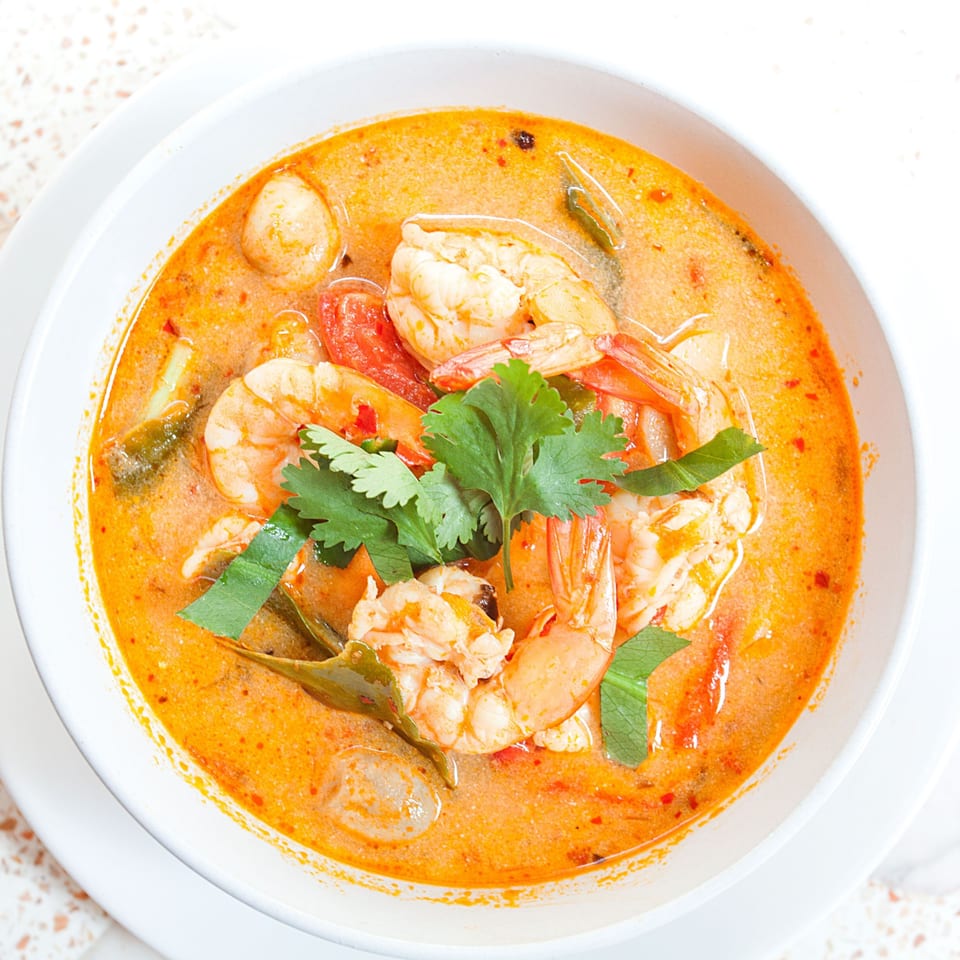 A vibrant orange bowl of Thai Tom Yum soup with shrimp, tomatoes, and fresh herbs.
