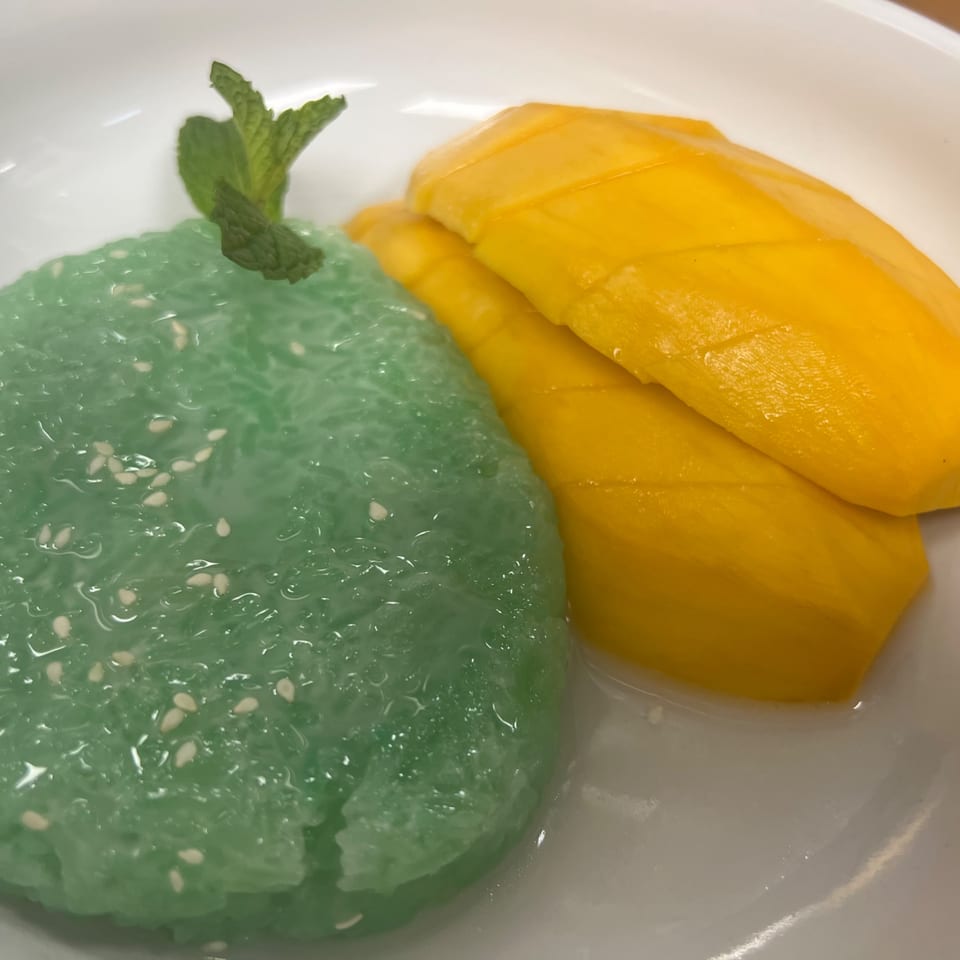 Green sticky rice with sesame seeds and sliced yellow mango, garnished with mint, on a white plate.