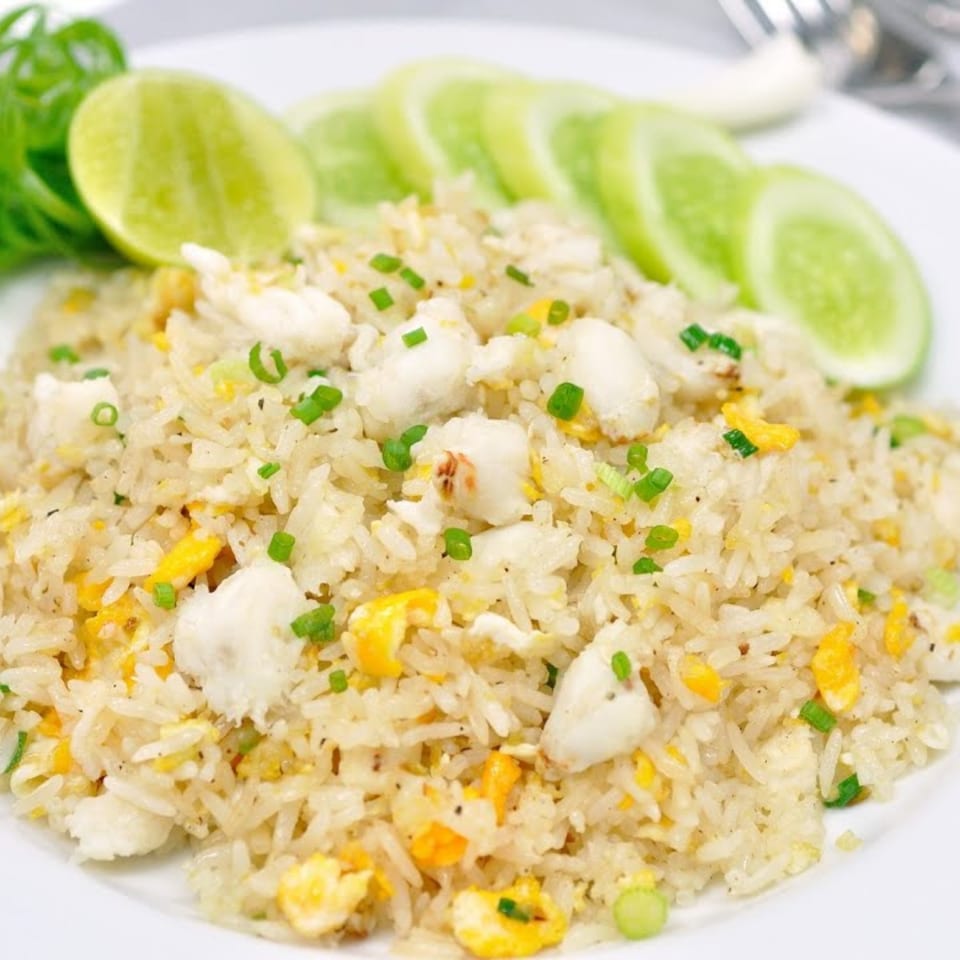 Thai crab fried rice with egg, green onions, lime wedges, and cucumber slices on a white plate.