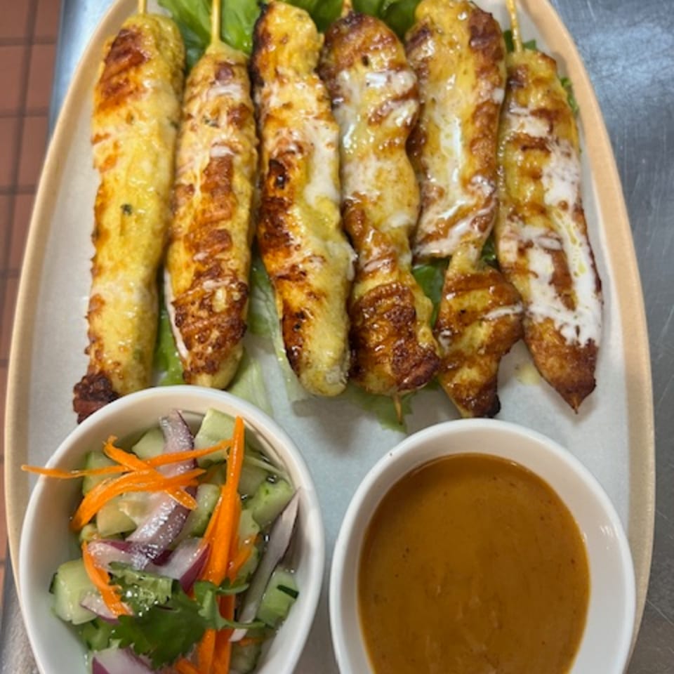 Thai chicken satay skewers with peanut sauce and cucumber salad on a beige platter.